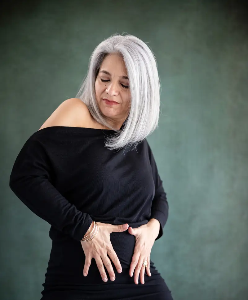 A woman with silver hair in black, hands resting at her waist, eyes lowered.