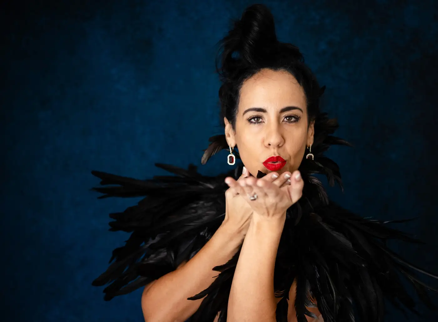 Woman in black feathers raising her hands toward her lips against a blue backdrop
